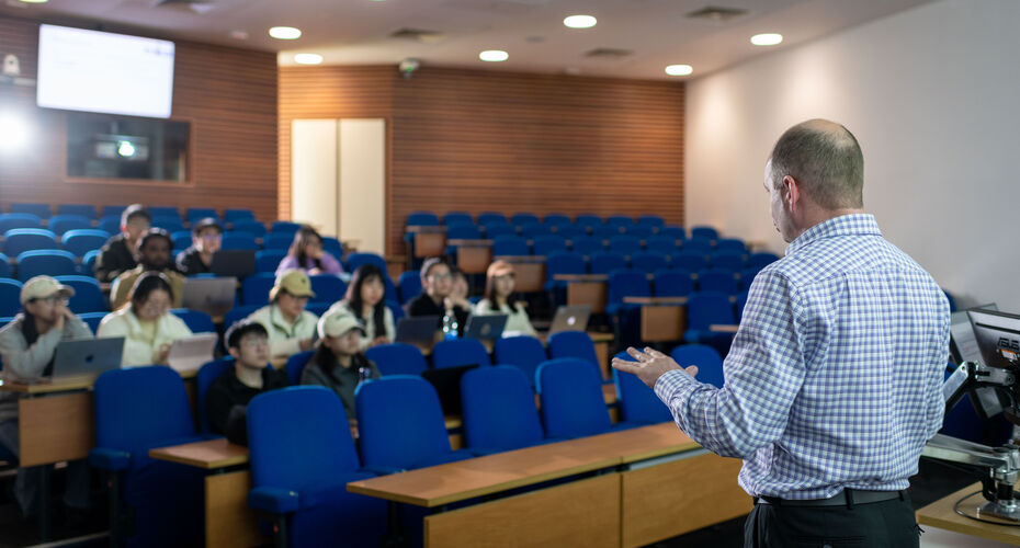 A academic delivers a lecture in an lecture theatre, engaging students seated at desks in front of him.