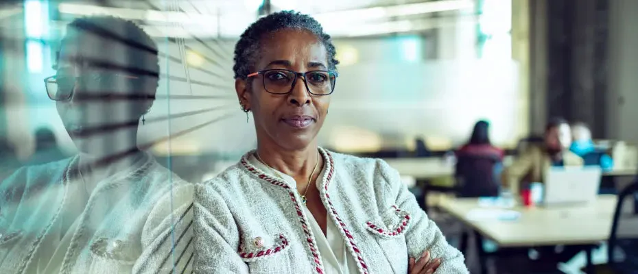 An older woman with glasses stands in front of a transparent glass wall, showcasing her thoughtful expression.