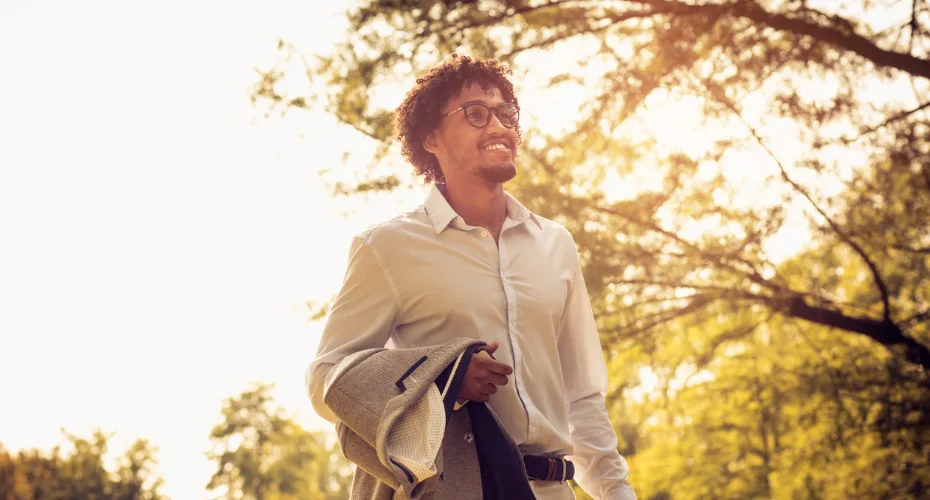 A man in a white shirt and tie walks through a park, surrounded by greenery and trees.