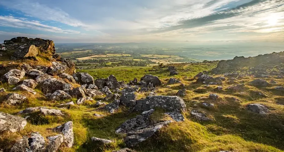 The sun shines brightly over a rocky hillside in Dartmoor, illuminating the rugged landscape.