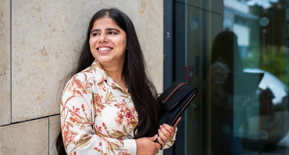 A smilingstudent holding a briefcase stands against a wall outside building one at the university of exeter business school.