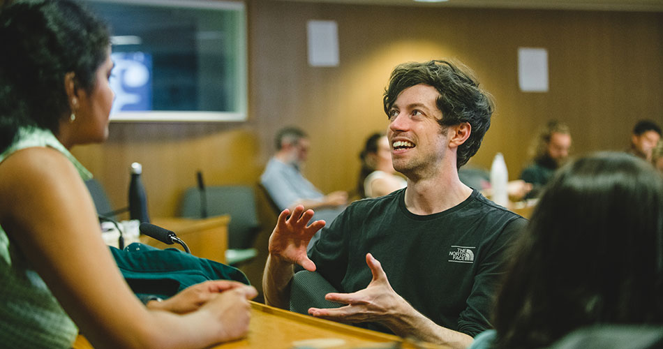 A man with curly hair and a black shirt sits at a table, engaged in conversation with a woman in a green top, both smiling and gesturing with their hands.